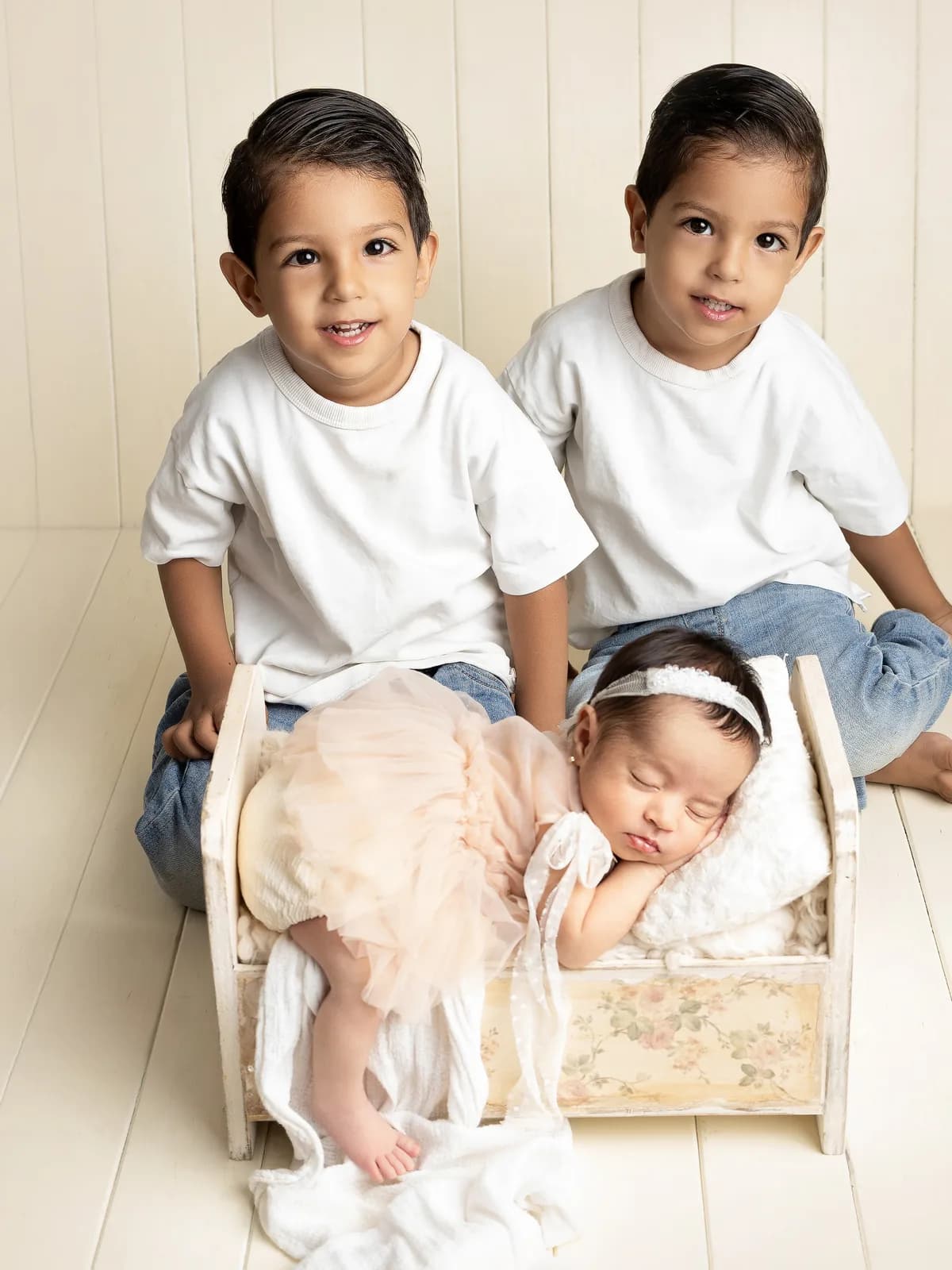 Dos hermanos mayores sentados junto a su hermana recién nacida en una cuna de madera, durante sesión newborn familiar en Querétaro